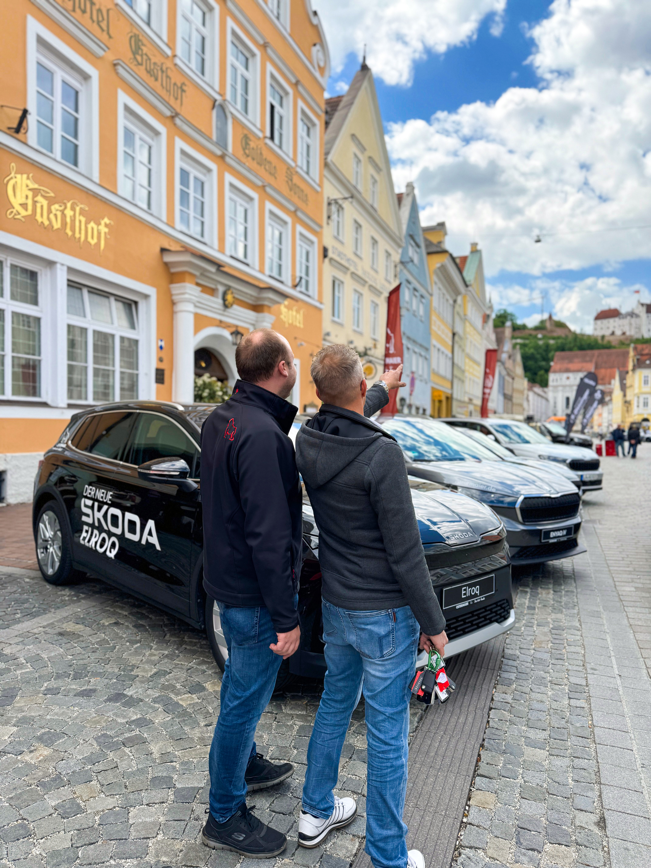 Zwei Männer betrachten Škoda-Autos bei einer Autoschau in der Altstadt. Einer zeigt auf bunte Häuser und eine Burg im Hintergrund. Links steht ein Škoda Elroq mit roter Beschriftung.