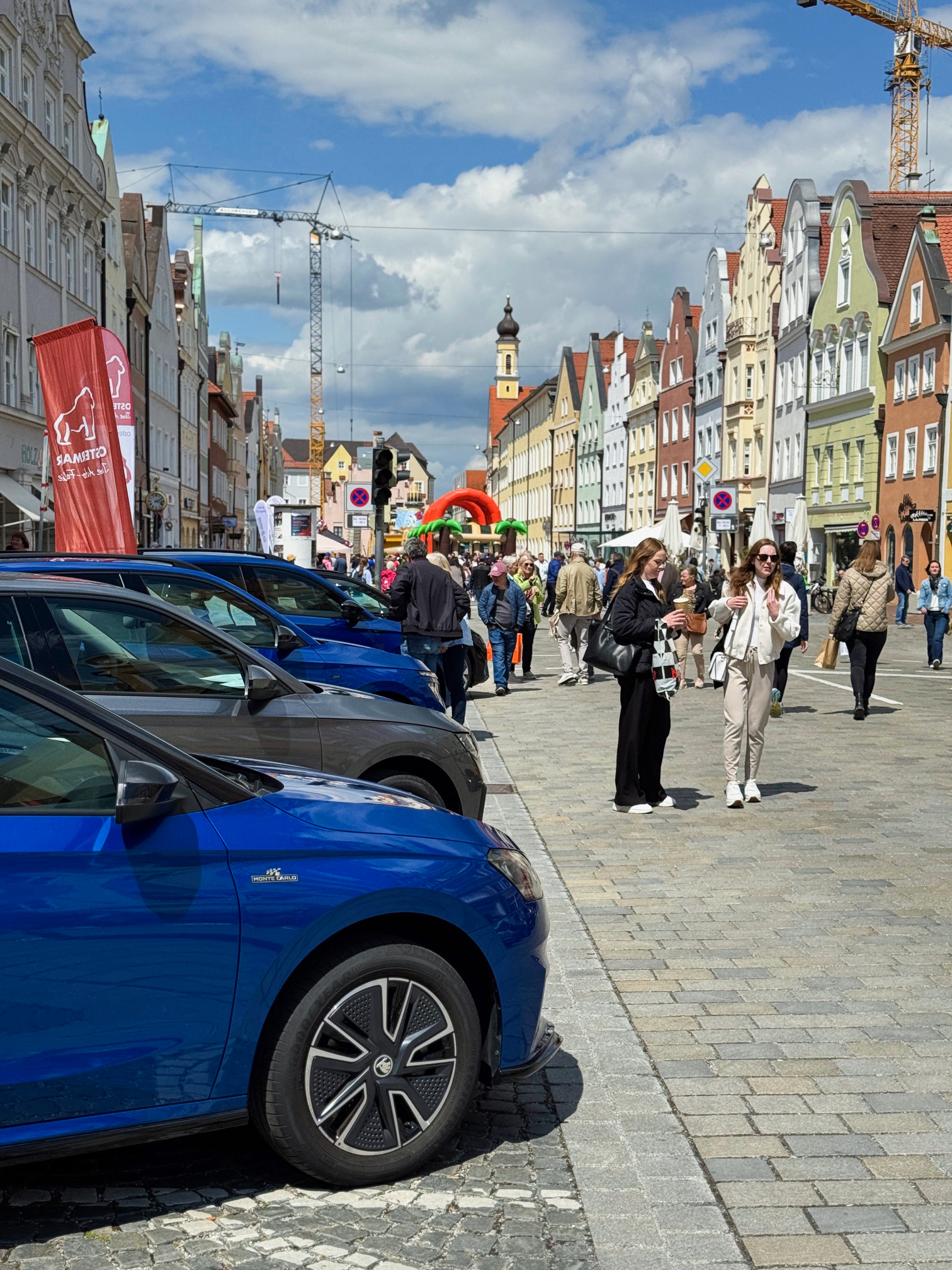 Menschen flanieren über eine belebte Altstadtstraße mit parkenden Škoda-Autos. Bunte Fassaden, ein Kirchturm, Kräne und eine Hüpfburg sind im Hintergrund zu sehen.